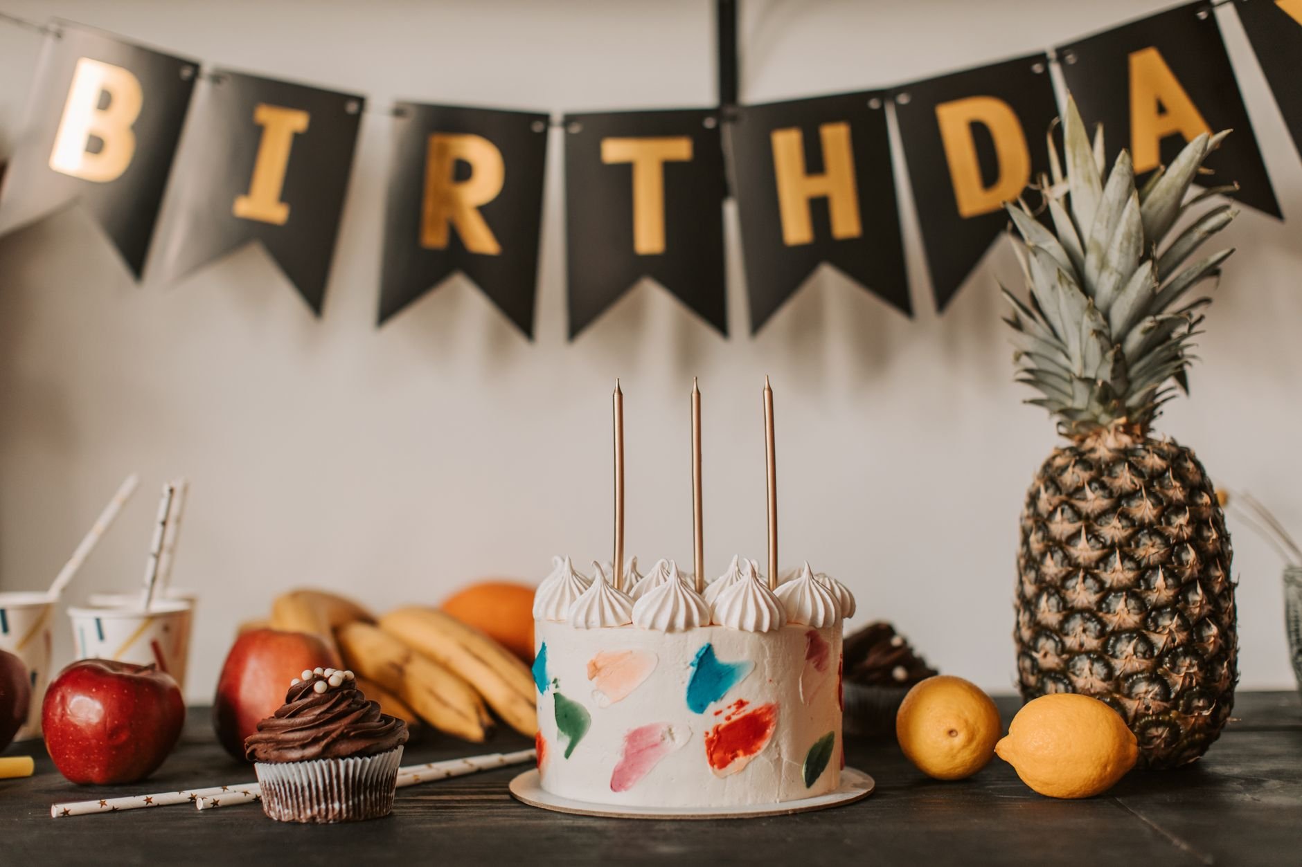 A colorful birthday cake with fruits and festive decorations on a table indoors.